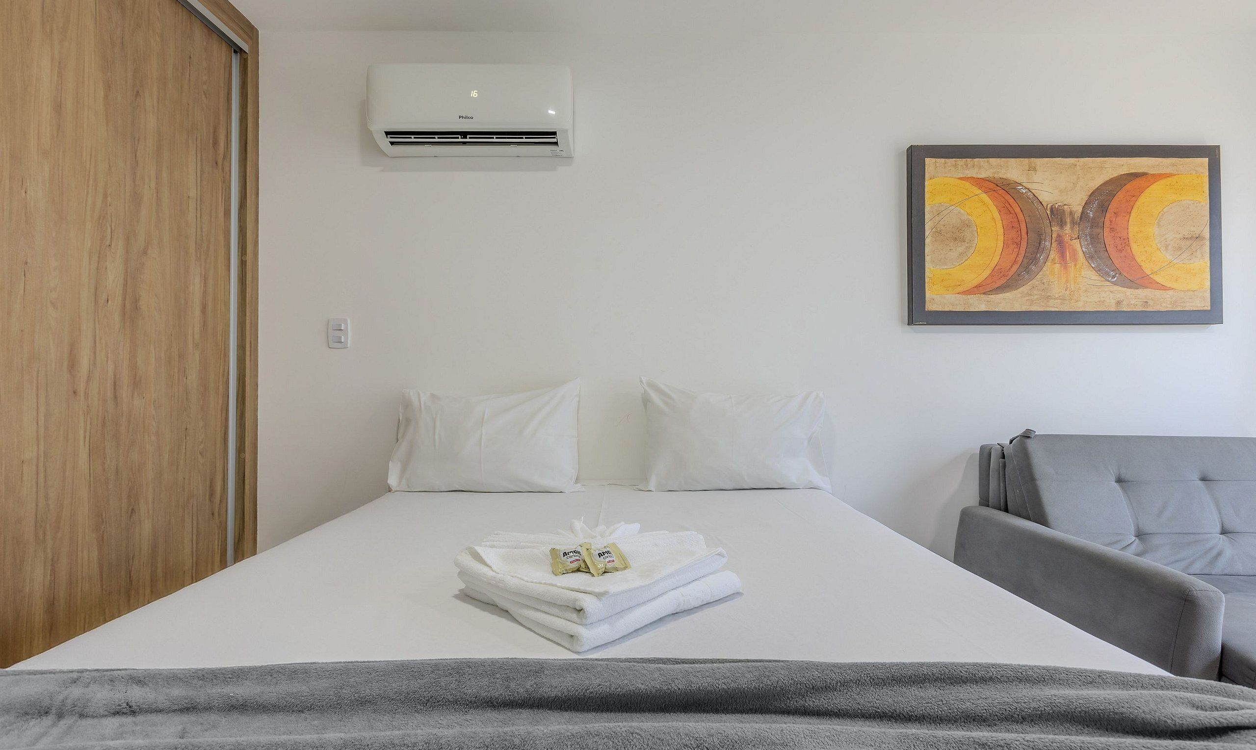 A hotel room with a bed, a gray armchair, an air conditioning unit, and a painting on the wall.