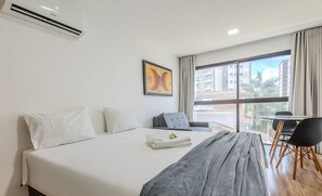 A hotel room with a bed, a gray chair, a small table, and a window with a view of buildings. - Studio With Pool in Pocos de Caldas Pgo412 (Poços de Caldas)