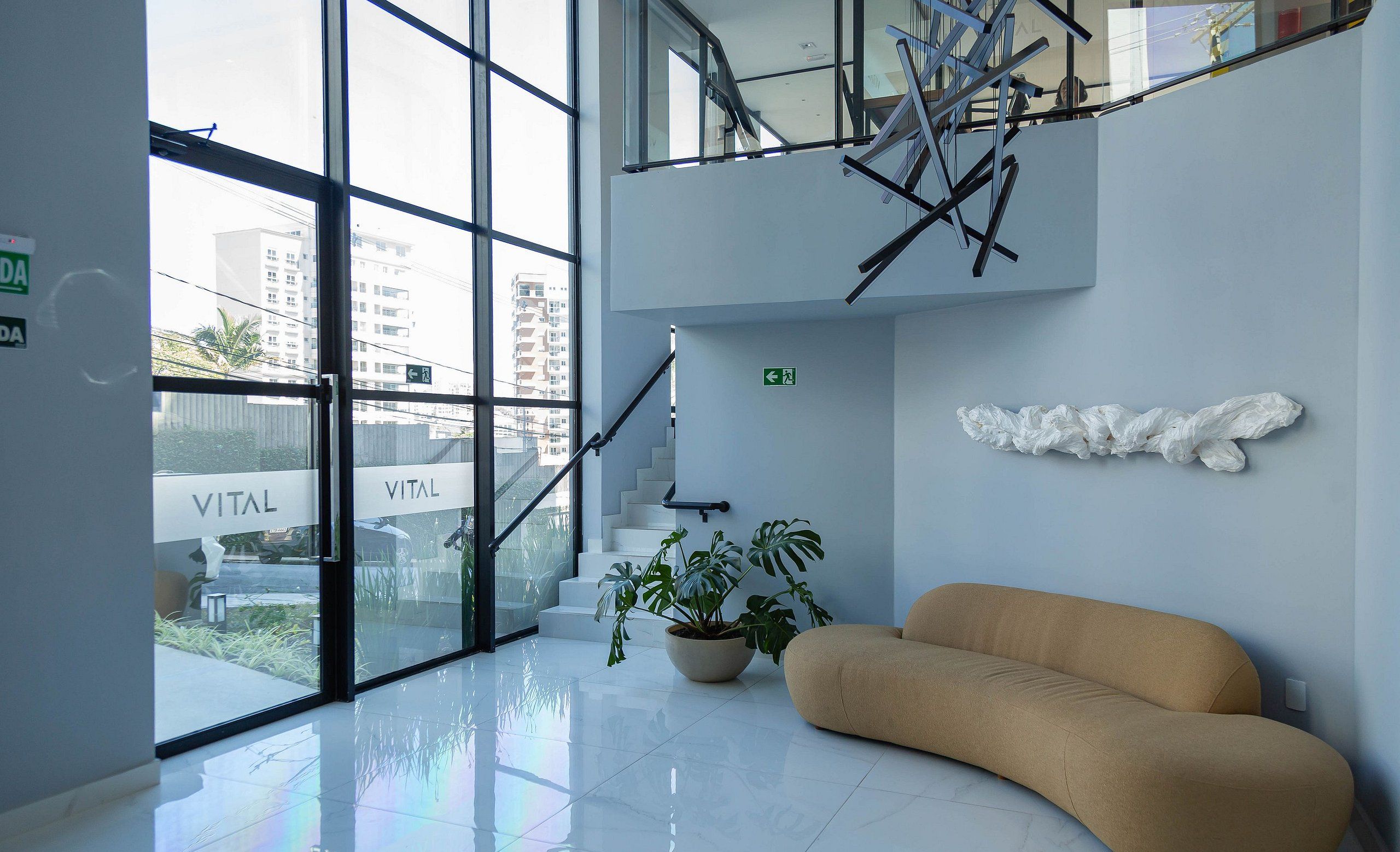 Un lobby moderno con entrada de vidrio, una escalera, una planta en maceta y un sofá color crema.