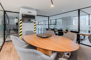 A modern meeting room with a round wooden table, grey upholstered chairs, and a glass partition with windows.