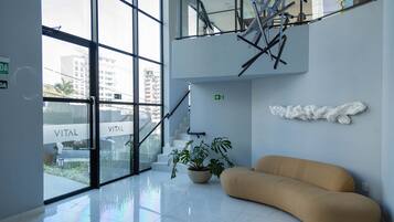 Un lobby moderno con entrada de vidrio, una escalera, una planta en maceta y un sofá color crema.