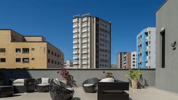 Terraza en la azotea con mobiliario de mimbre y plantas en macetas, rodeada de edificios modernos.