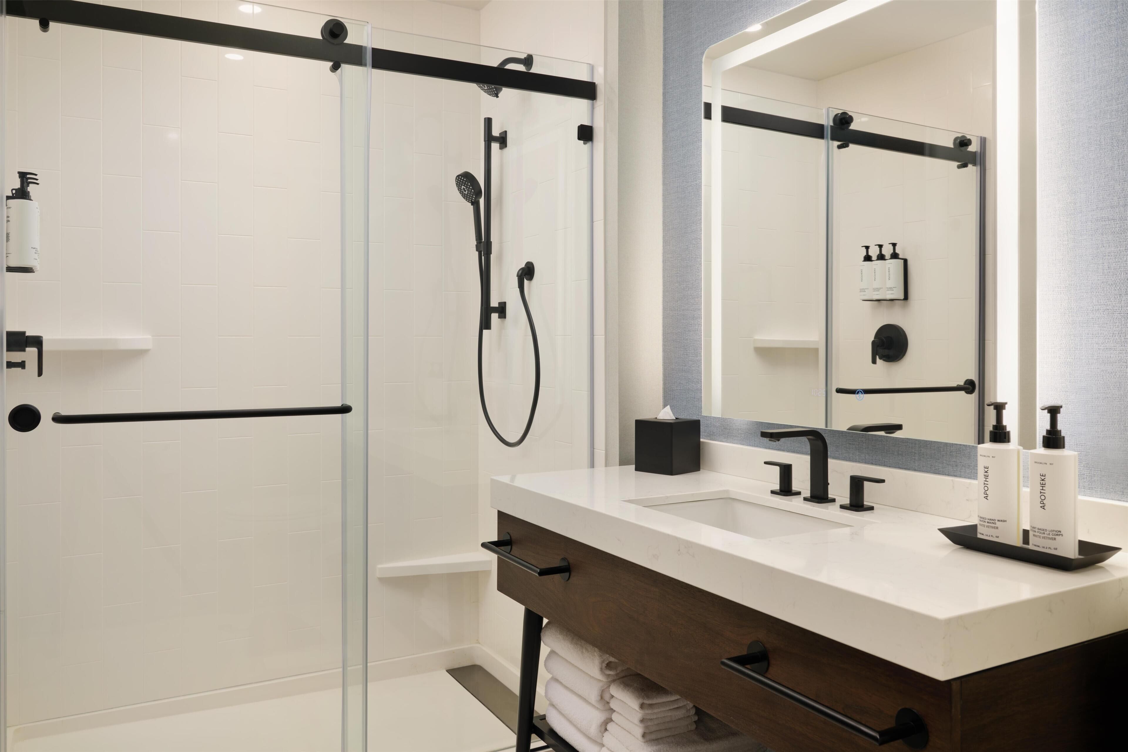 A modern bathroom with a glass shower enclosure, a white sink with a dark countertop, and a large mirror.