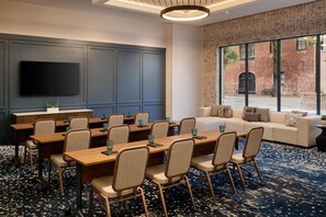 Une salle de conférence équipée d’un grand téléviseur à écran plat, de plusieurs tables avec des chaises, d’un canapé et d’une grande fenêtre donnant sur un bâtiment en briques.