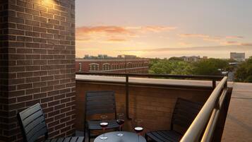 A balcony with brick walls, two tables, and three chairs, overlooking a cityscape during sunset.