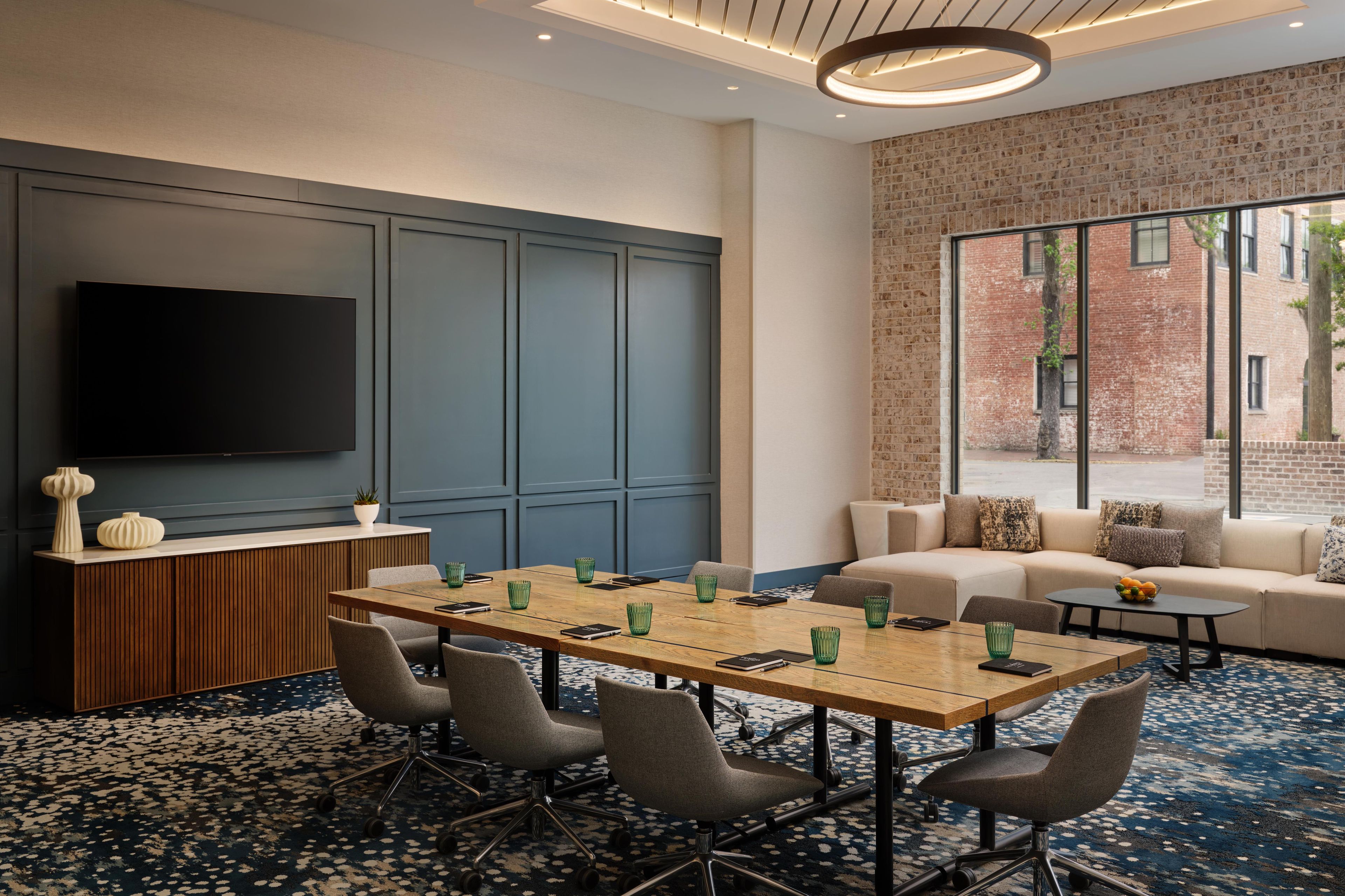 A conference room with a long table, chairs, a TV, a sofa, and a large window with a brick wall view.