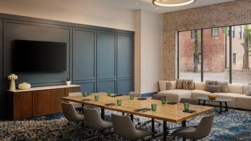 A conference room with a long table, chairs, a TV, a sofa, and a large window with a brick wall view.