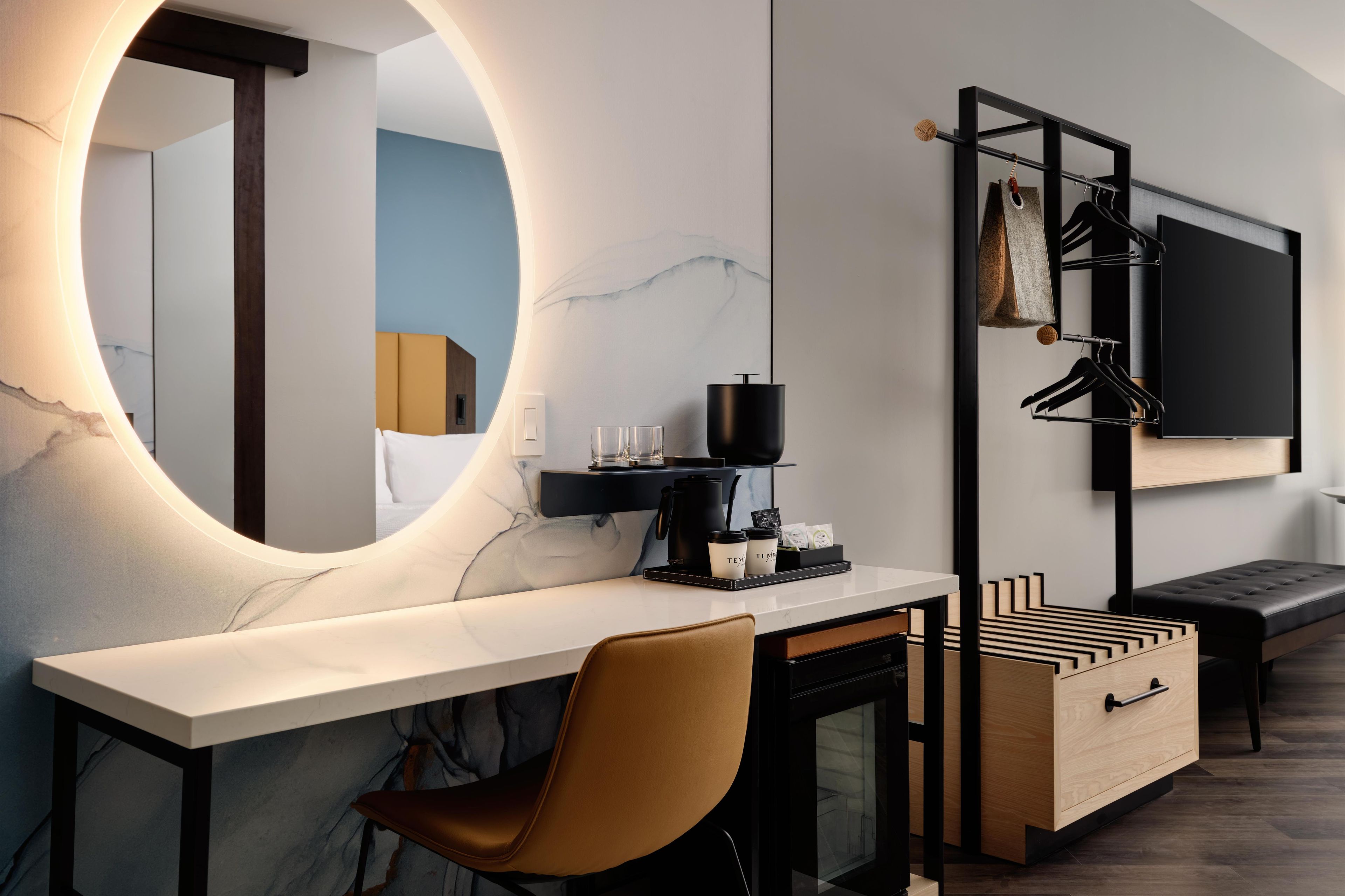 A modern hotel room with a marble desk, a large mirror, a black TV mounted on the wall, a wooden bench, and a black coat rack with hangers.