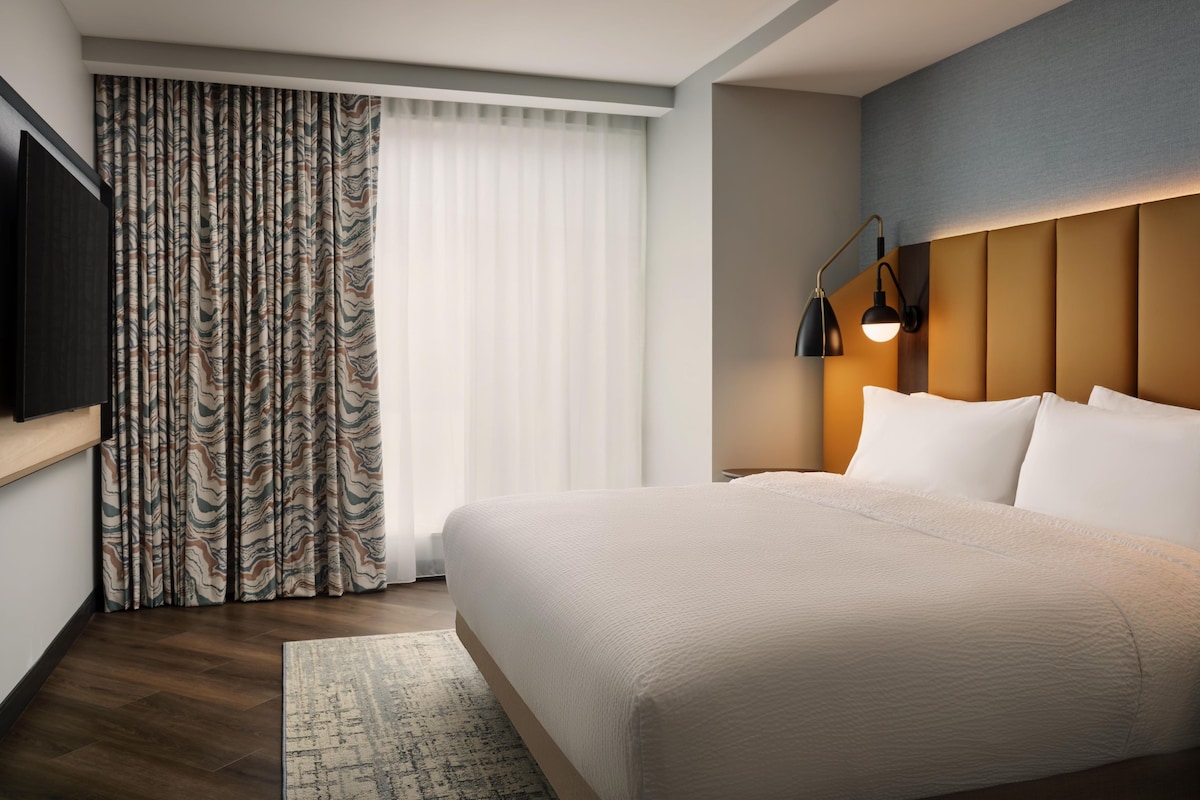 Ein Hotelzimmer mit einem Bett, einem Fernseher, einem Nachttisch, einer Lampe und einem Fenster mit Vorhängen.