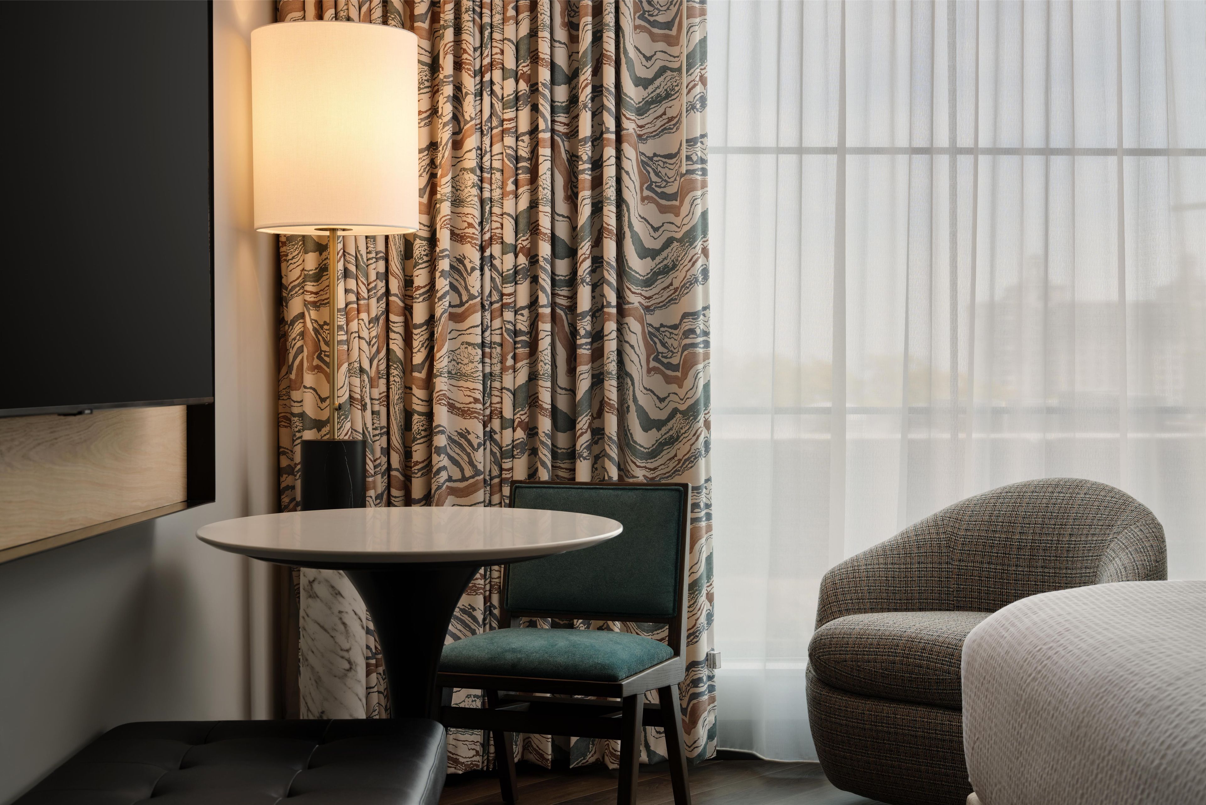 Een hotelkamer met een ronde tafel, een groene stoel, een bank, een lamp en gordijnen met een patroon.