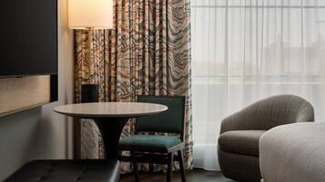 Een hotelkamer met een ronde tafel, een groene stoel, een bank, een lamp en gordijnen met een patroon.