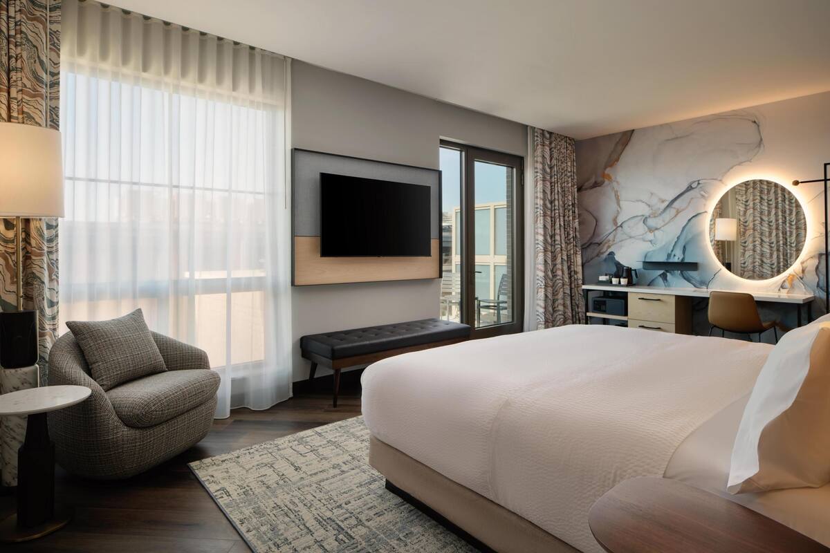 A hotel room with a large bed, a flat-screen TV mounted on the wall, a desk with a chair, a round mirror, and a view of the city through the window.