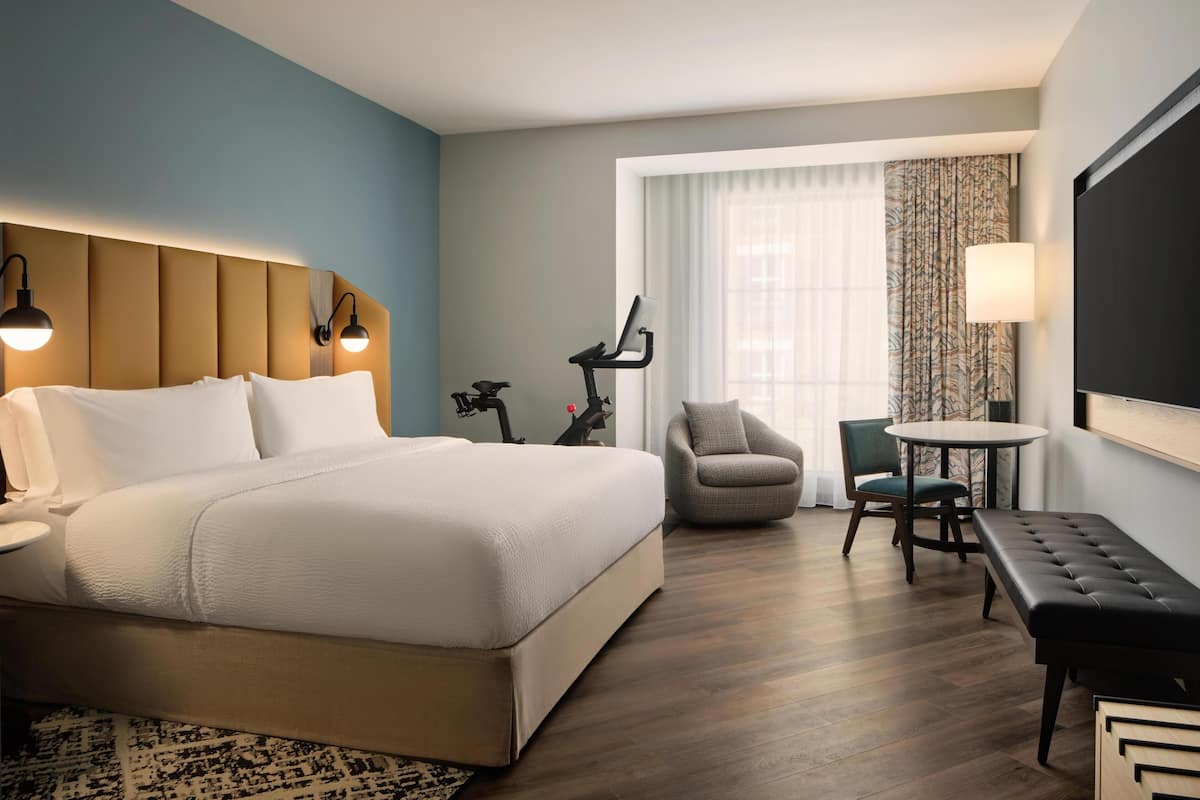 A hotel room with a large bed, a desk with a chair, a treadmill, and a TV.