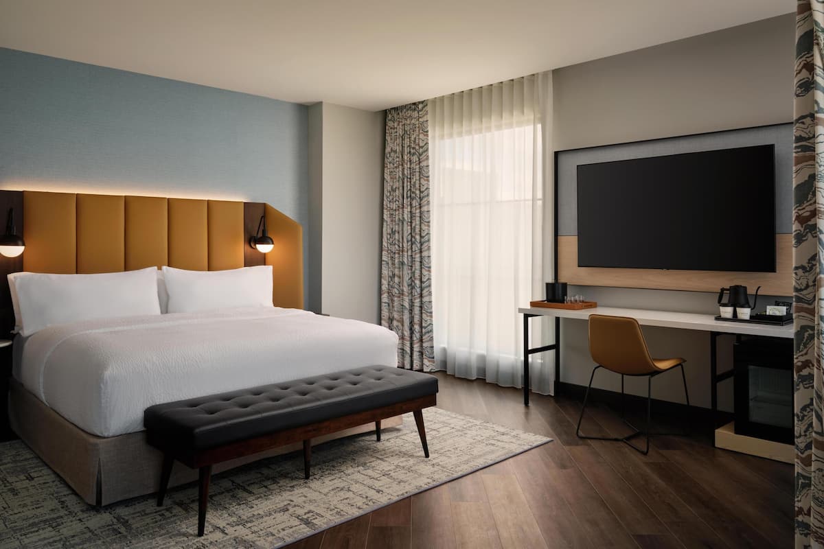 A hotel room with a bed, a desk, a chair, a large TV, and a bench.