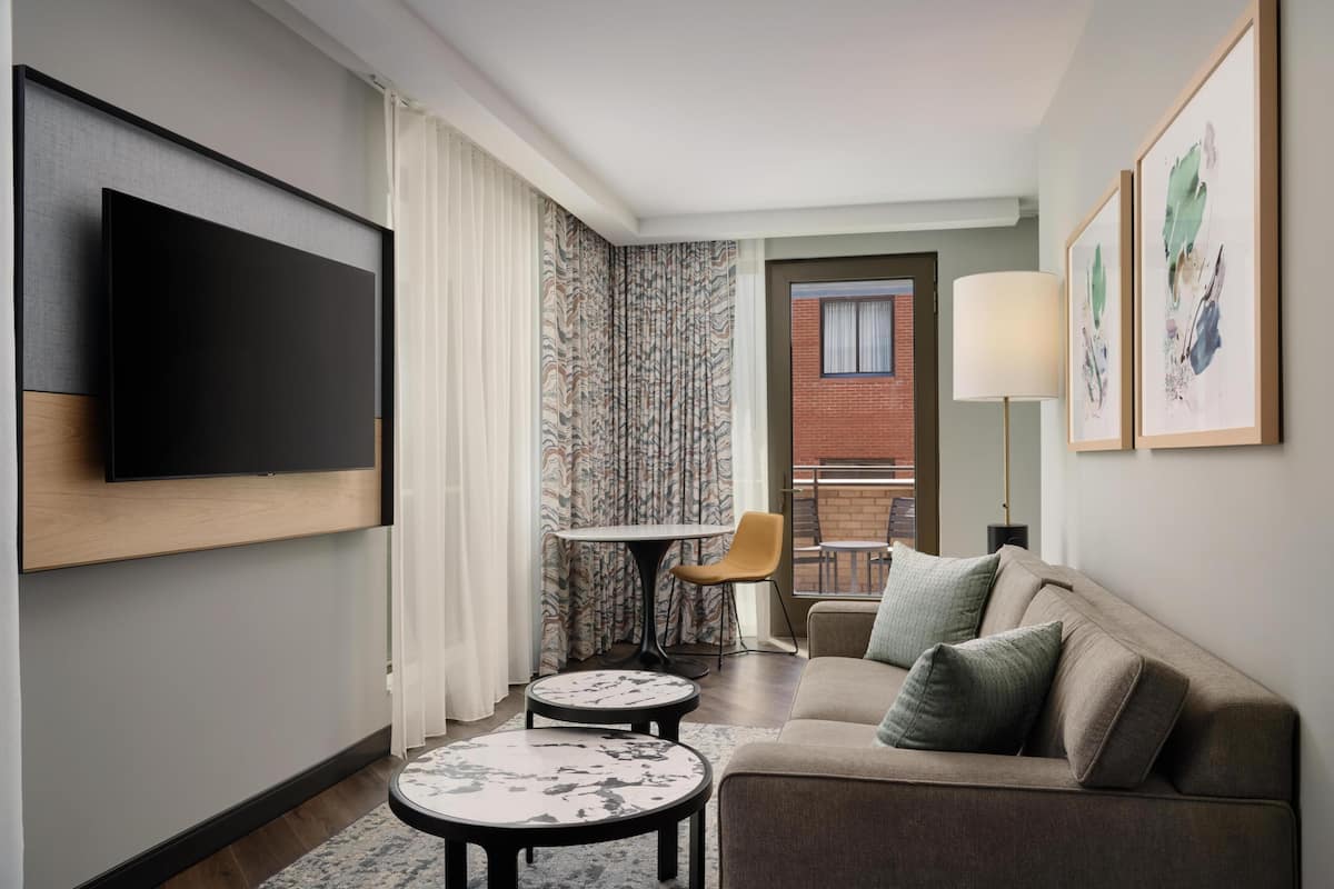 A modern hotel room with a flat-screen TV, a sofa, a small round table, a chair, and a balcony with a view.