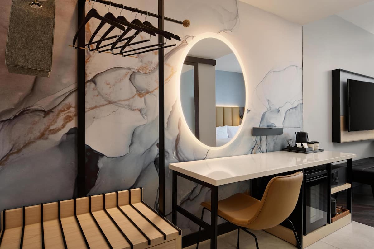 A modern bathroom with a marble wall, a round mirror with lighting, a towel rack, a wooden bench, and a black chair.