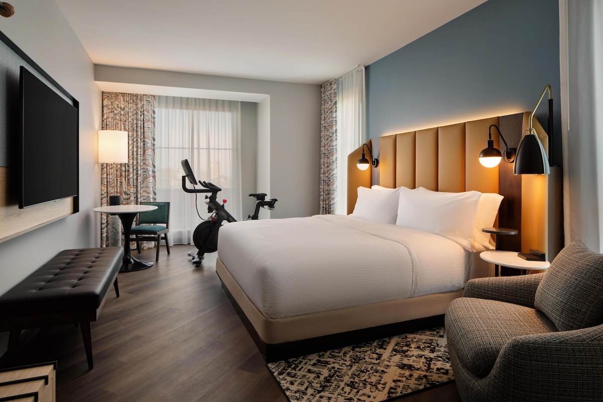 A hotel room with a large bed, a desk, a chair, a television, and a wall-mounted lamp.
