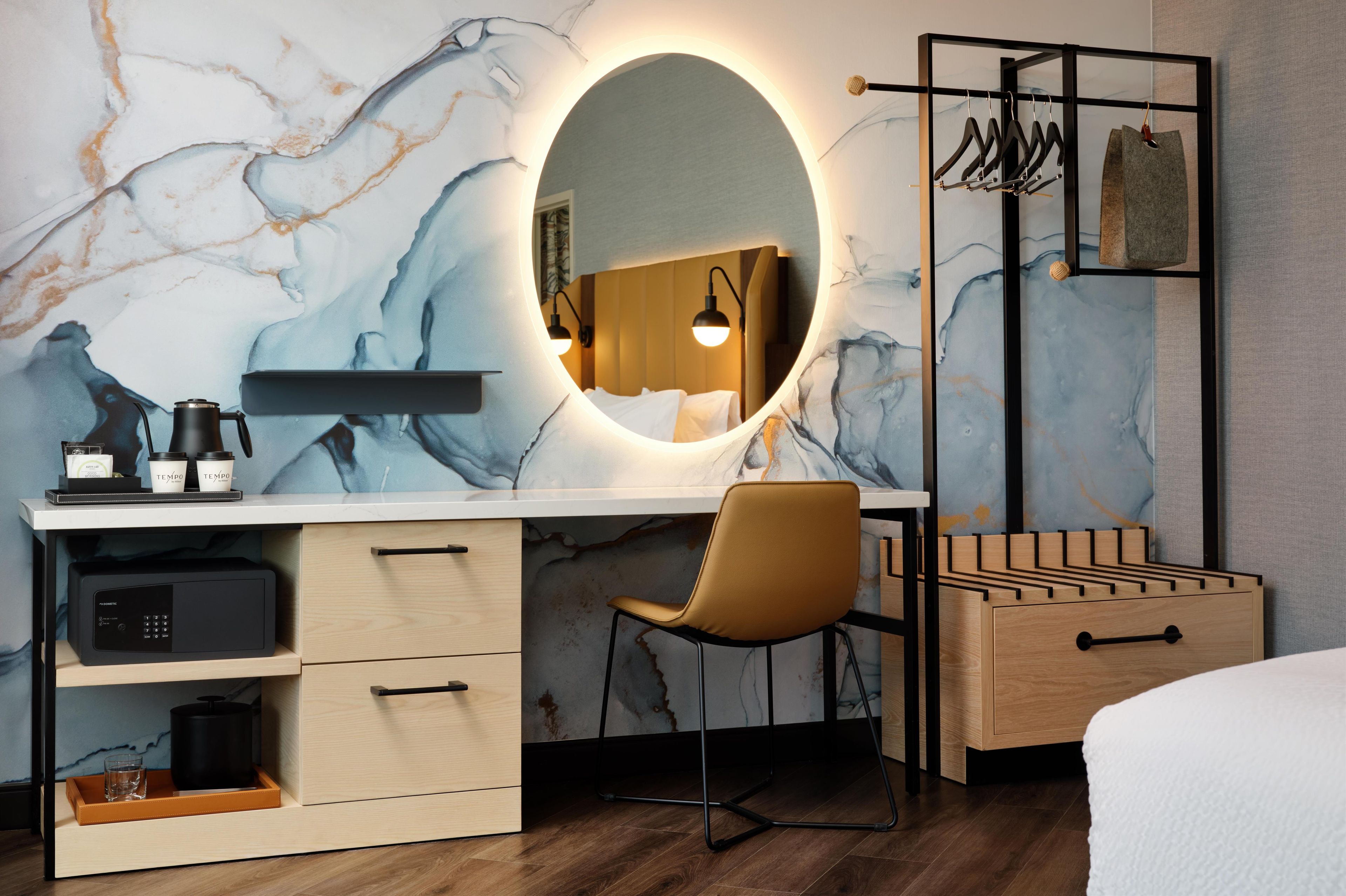 A modern bedroom with a marble wall, a round mirror, a wooden desk, a chair, a bed, and a hanging clothes rack.