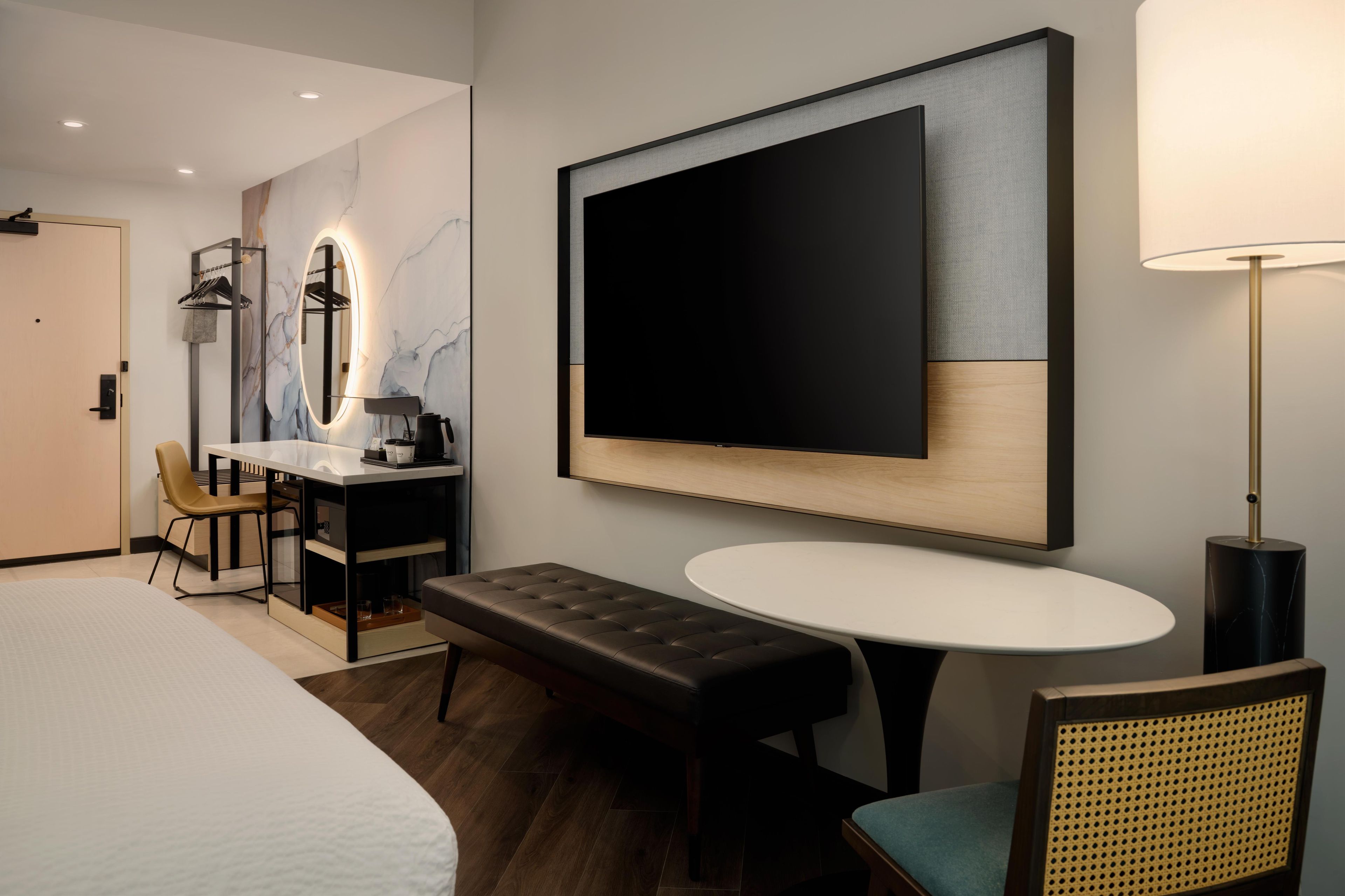 Ein Hotelzimmer mit einem Bett, einem Schreibtisch mit Stuhl, einem runden Tisch, einer Bank, einem an der Wand befestigten Fernseher und einer Lampe.