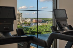 Une salle de sport équipée de matériel d’exercice et offrant une vue sur le paysage urbain grâce à de grandes fenêtres.