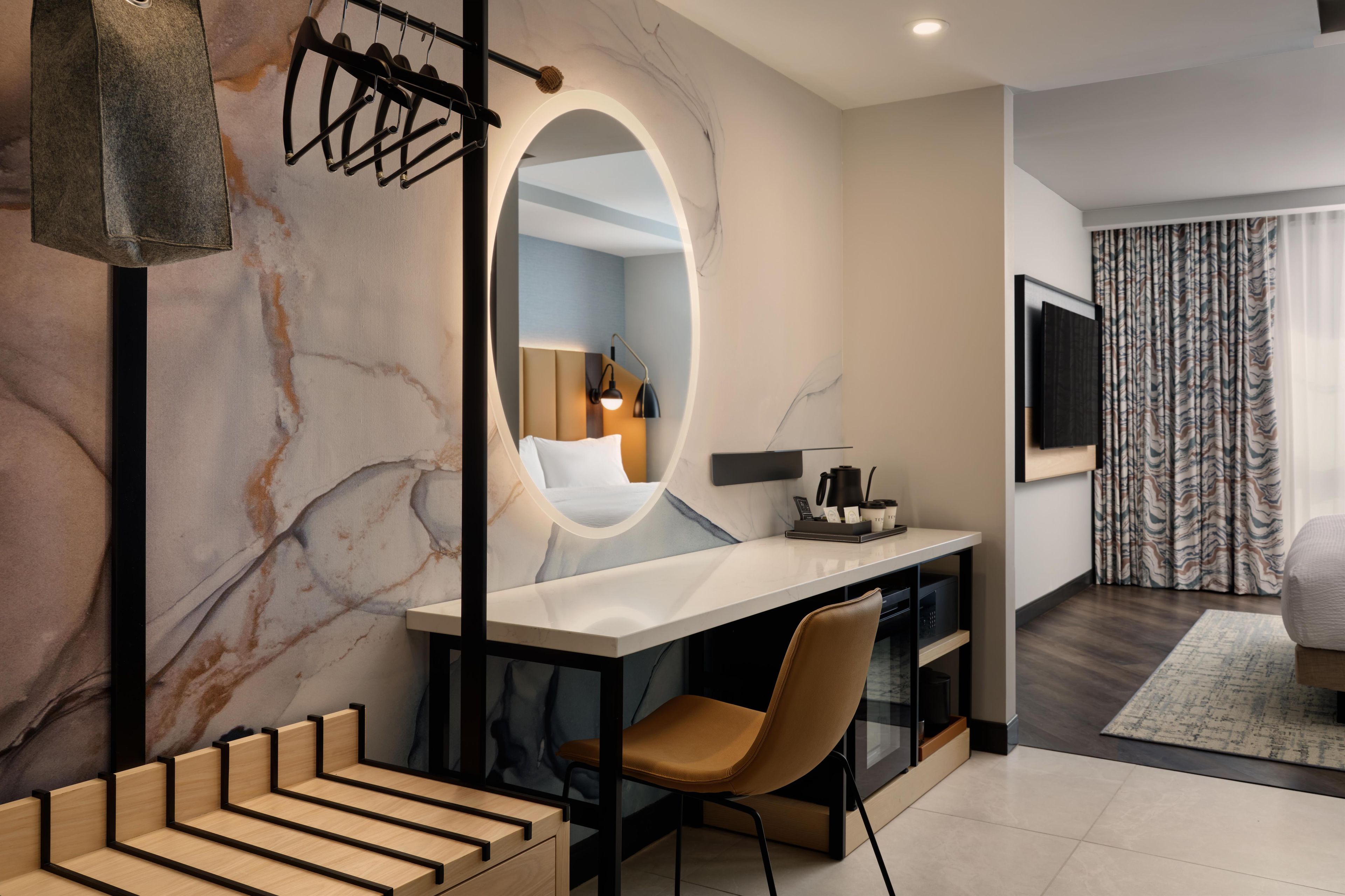 A modern hotel room with a marble wall, a large mirror, a desk with a chair, a TV mounted on the wall, and a patterned curtain.