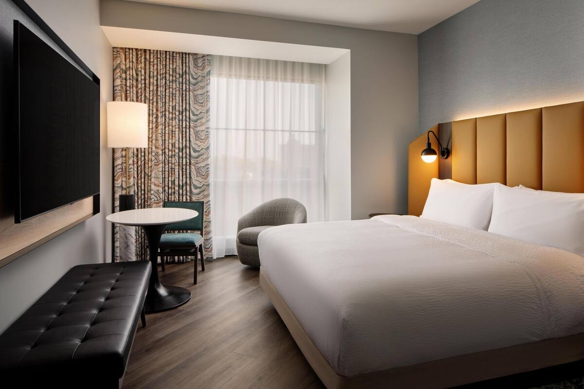 A hotel room with a bed, a TV, a round table, a bench, a chair, and a window with curtains.