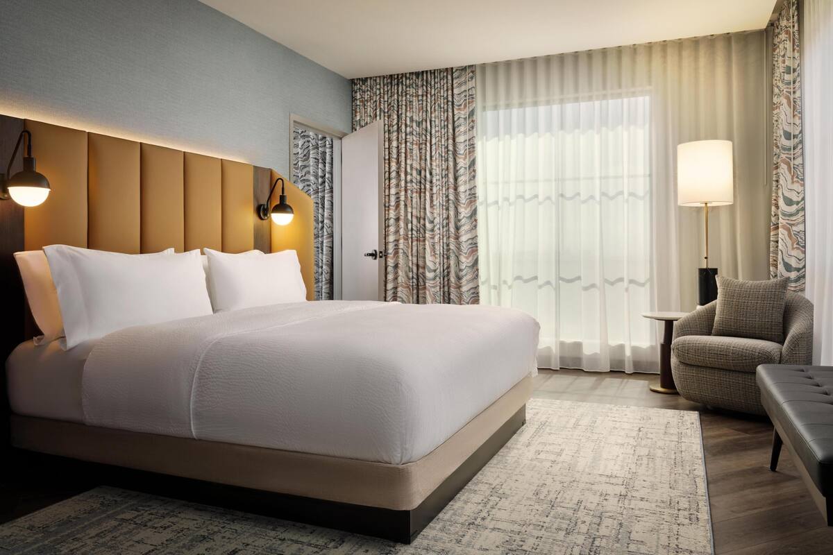 A hotel room with a large bed, a chair, a small table, and a lamp.