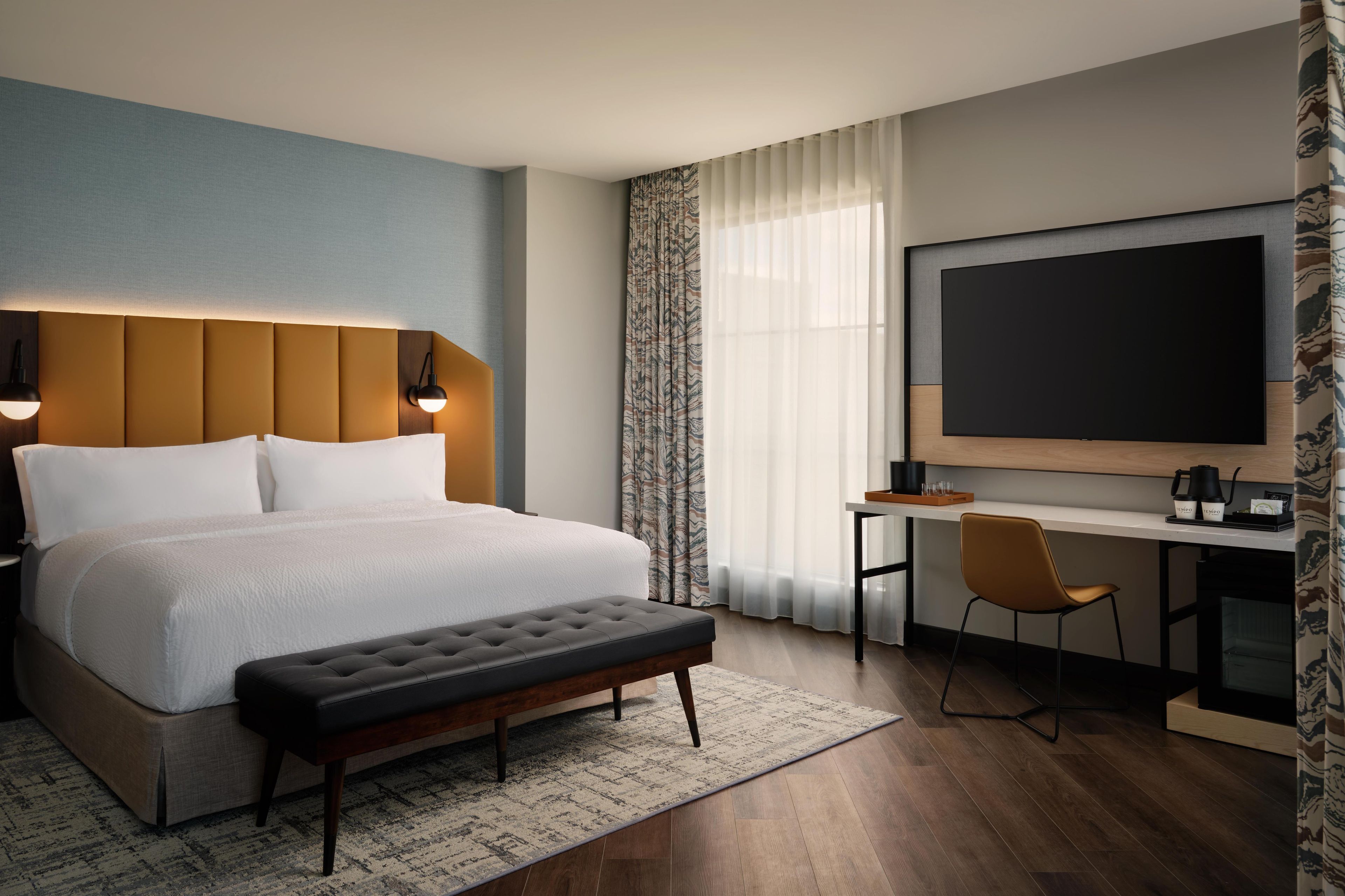 A hotel room with a bed, a desk, a chair, a large TV, and a bench.