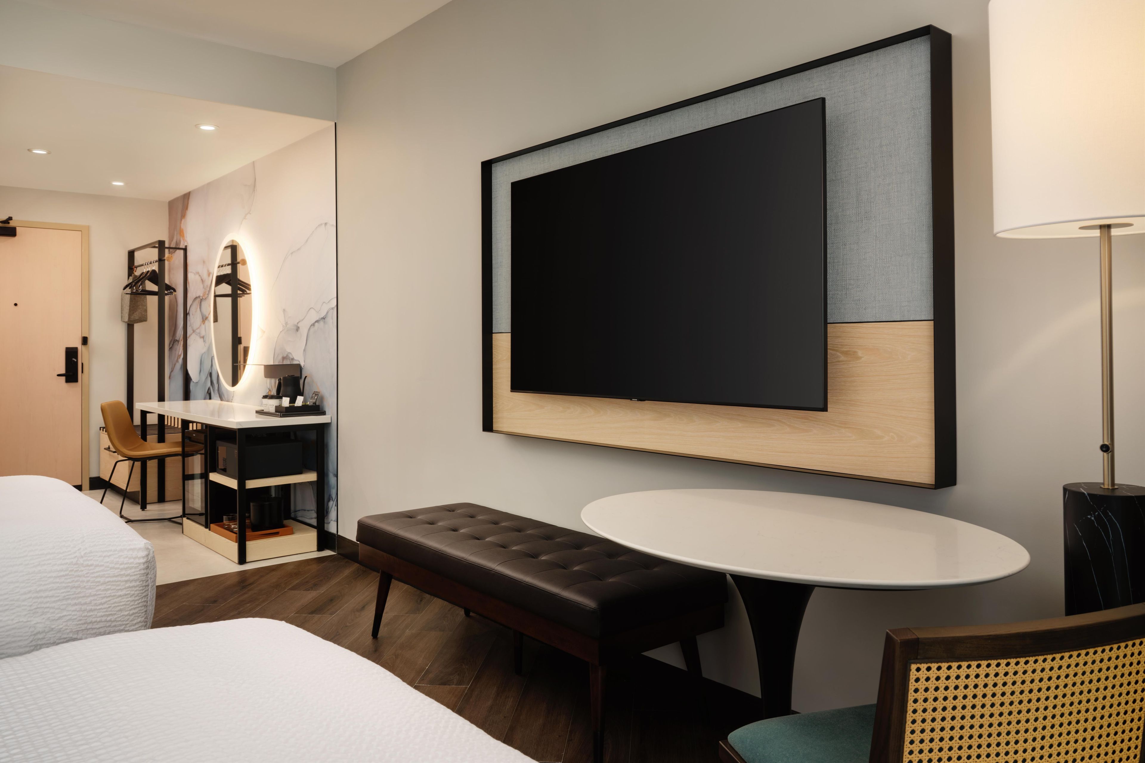 A hotel room with a bed, a round table, a bench, a desk with a chair, a mirror, and a large framed artwork on the wall.