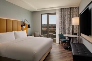 Habitación de hotel con una cama grande, un escritorio con una silla, una mesita, un sofá, un televisor y un balcón con vistas.