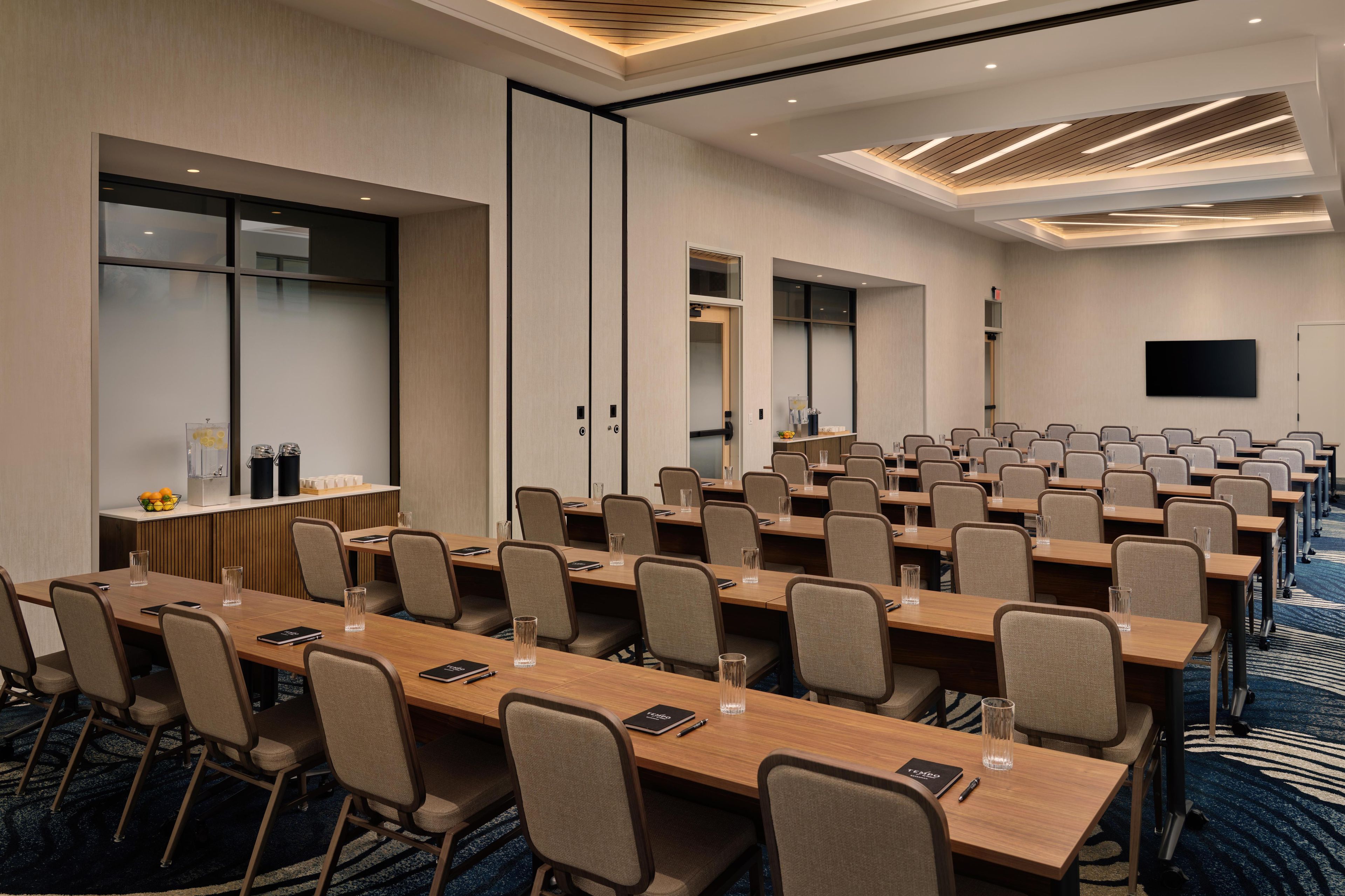 A conference room with long tables, chairs, a flat-screen TV, and a large window.