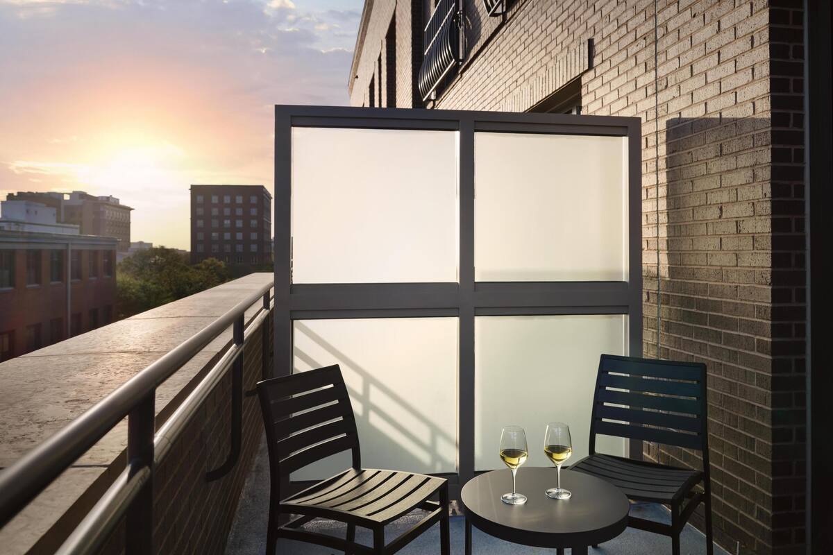 A balcony with two chairs and a table, two glasses of wine, and a view of buildings and a sunset.