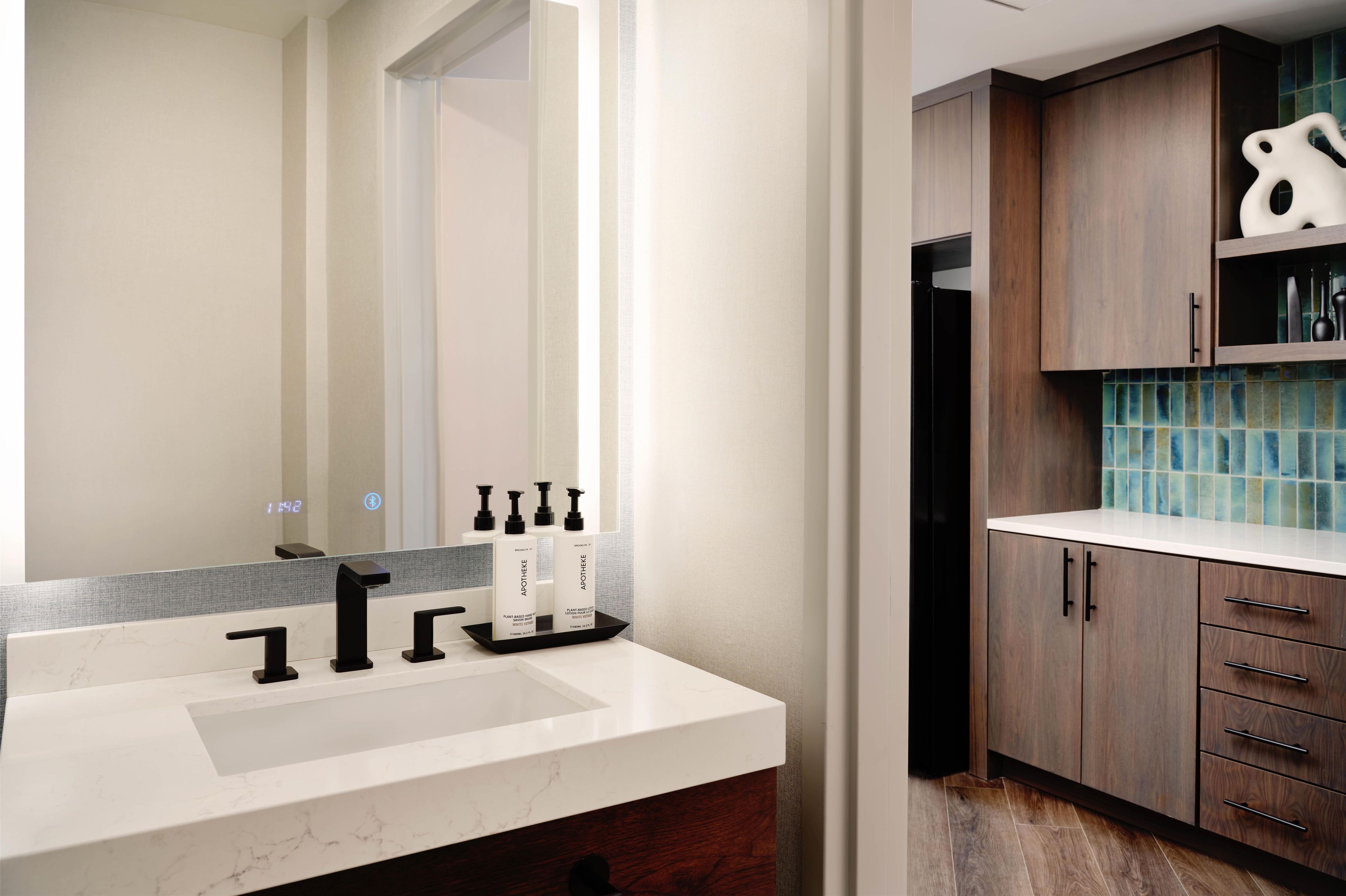 Un baño moderno con un lavamanos blanco, grifo negro y un espejo que refleja la habitación. Hay armarios de madera con un salpicadero de azulejos azules y una puerta negra.
