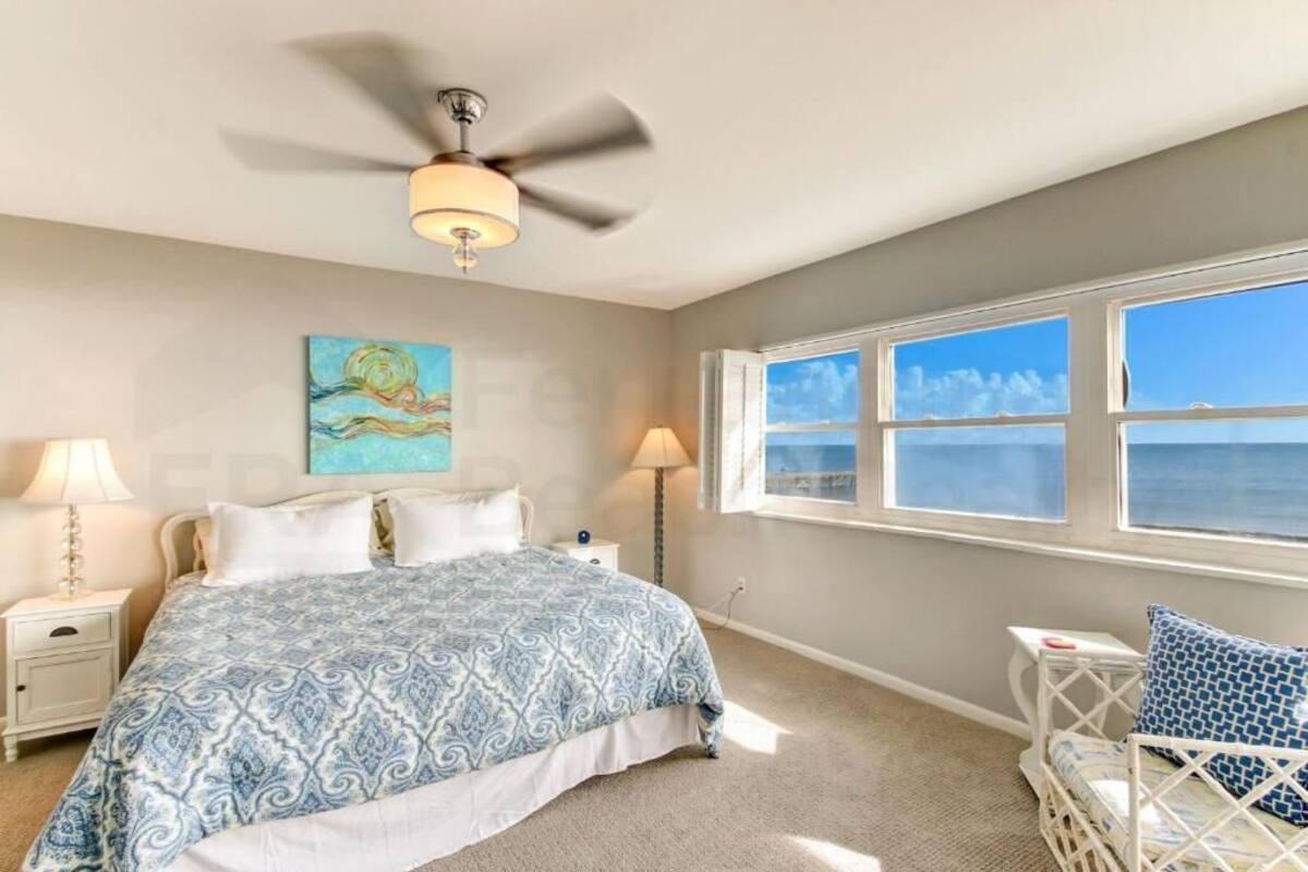 A bedroom with a bed, two bedside tables, a ceiling fan, a window with a view of the sea, and a painting on the wall.