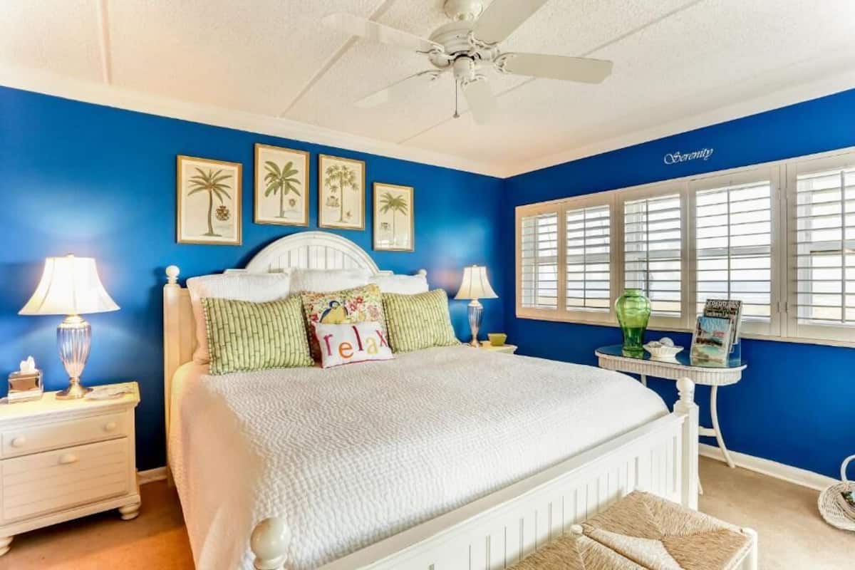 A bedroom with a white bed, blue walls, and a window with shutters.