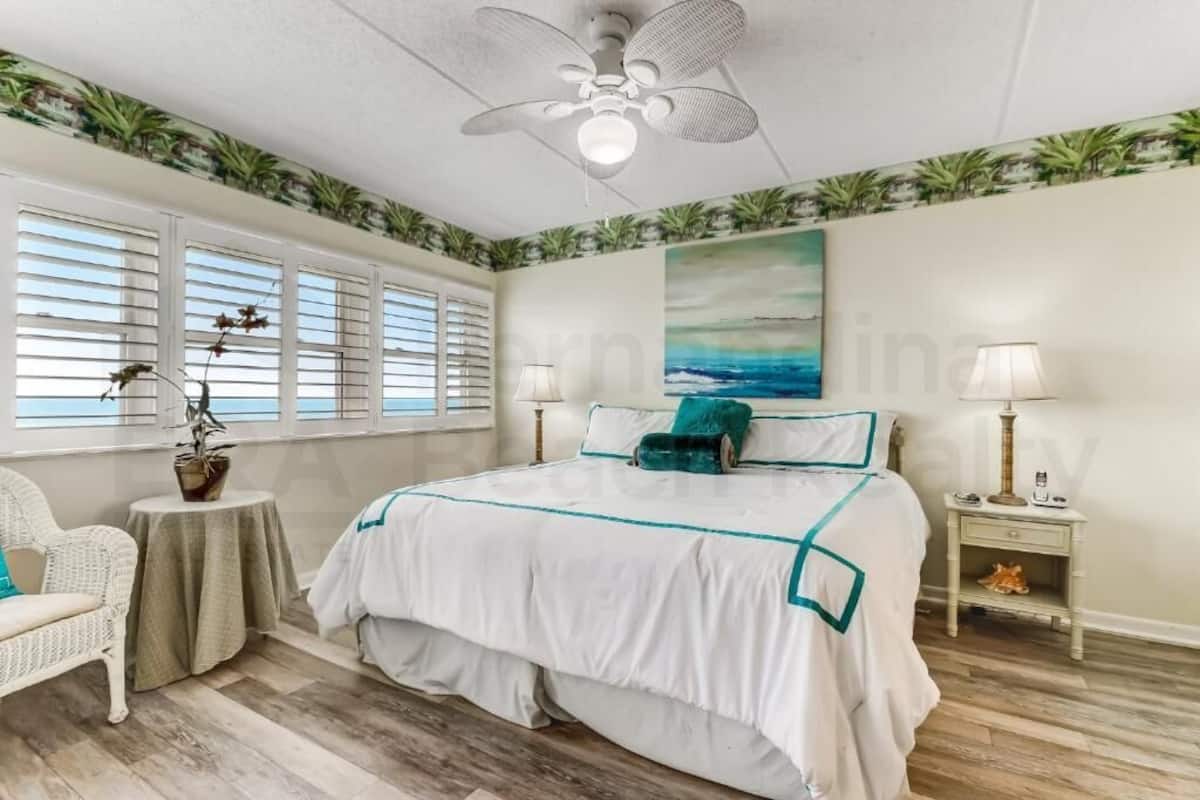 A bedroom with a bed, two bedside tables, a chair, a ceiling fan, and a window with blinds.