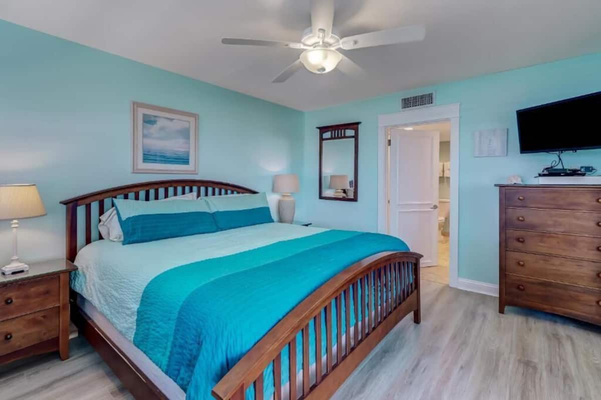 A bedroom with a bed, nightstands, a dresser, a ceiling fan, a TV, and a door.