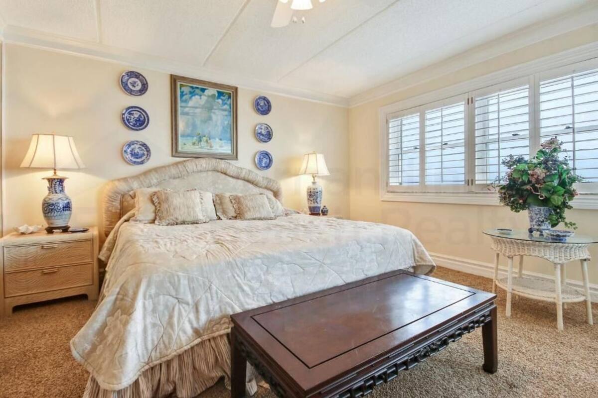 A bedroom with a large bed, two bedside tables with lamps, a small table with a plant, and a window with blinds.