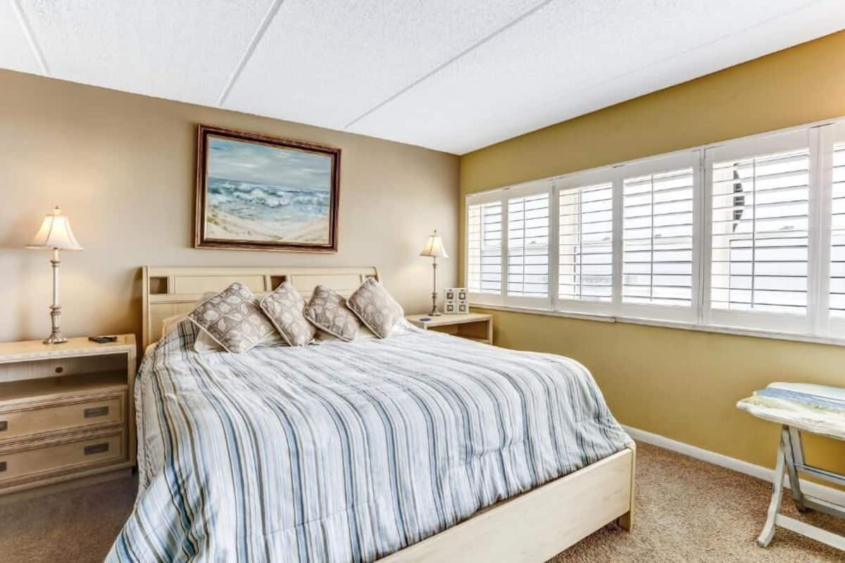 A bedroom with a bed, bedside tables, lamps, a painting, and a window with blinds.