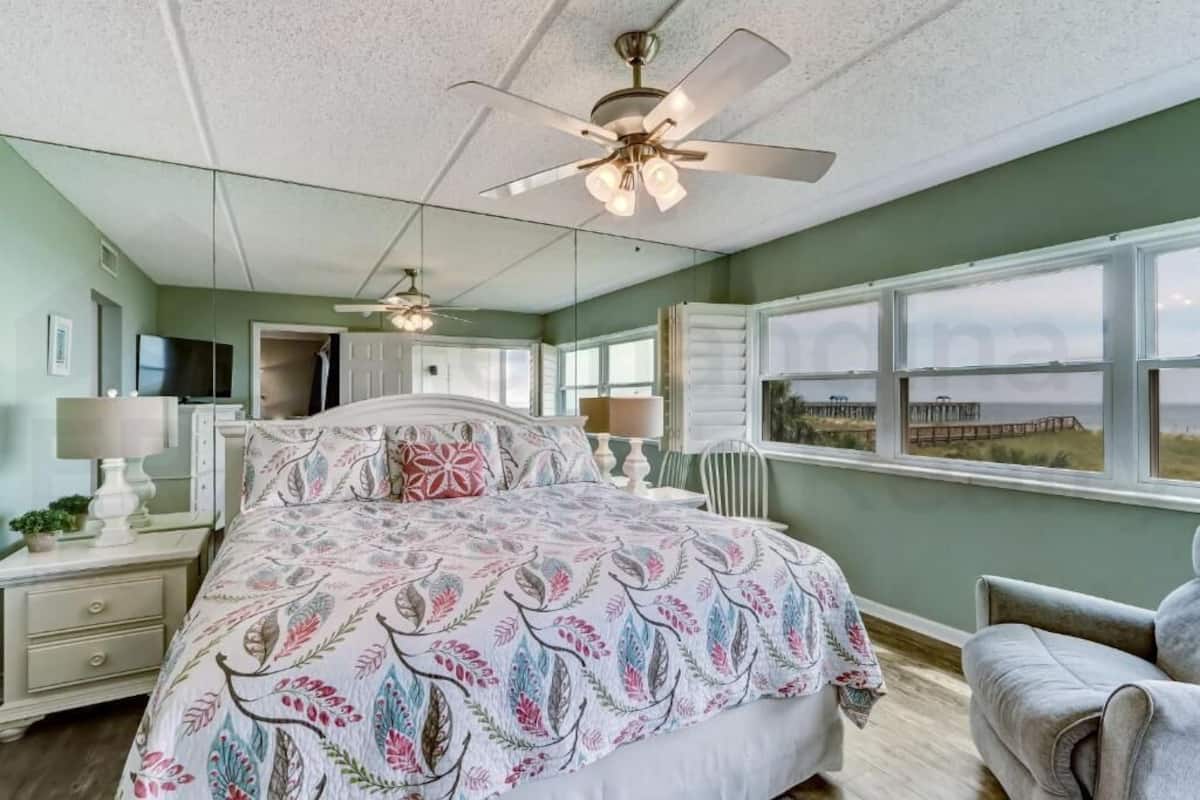 A bedroom with a bed, bedside tables, a ceiling fan, a window with a view, and a chair.