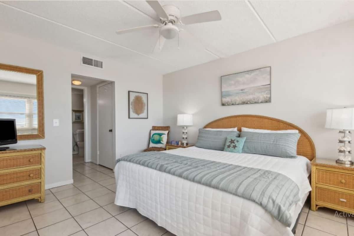 A bedroom with a bed, a television, a ceiling fan, a nightstand, and a wall-mounted lamp.