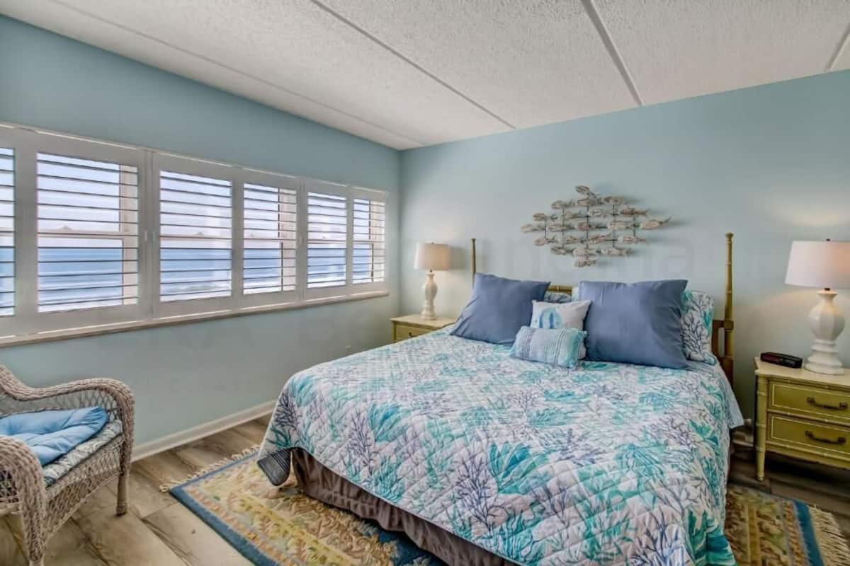 A bedroom with a large bed, two bedside tables, a chair, a window with blinds, and a decorative wall piece.