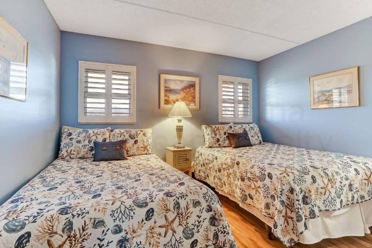 A bedroom with two beds, a nightstand, a lamp, and two windows with shutters.