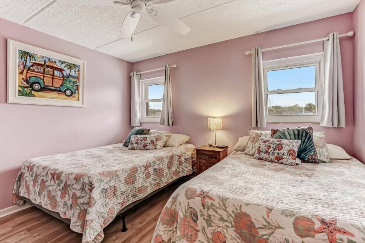 A bedroom with two beds, a ceiling fan, a window with curtains, a nightstand, and a painting on the wall.