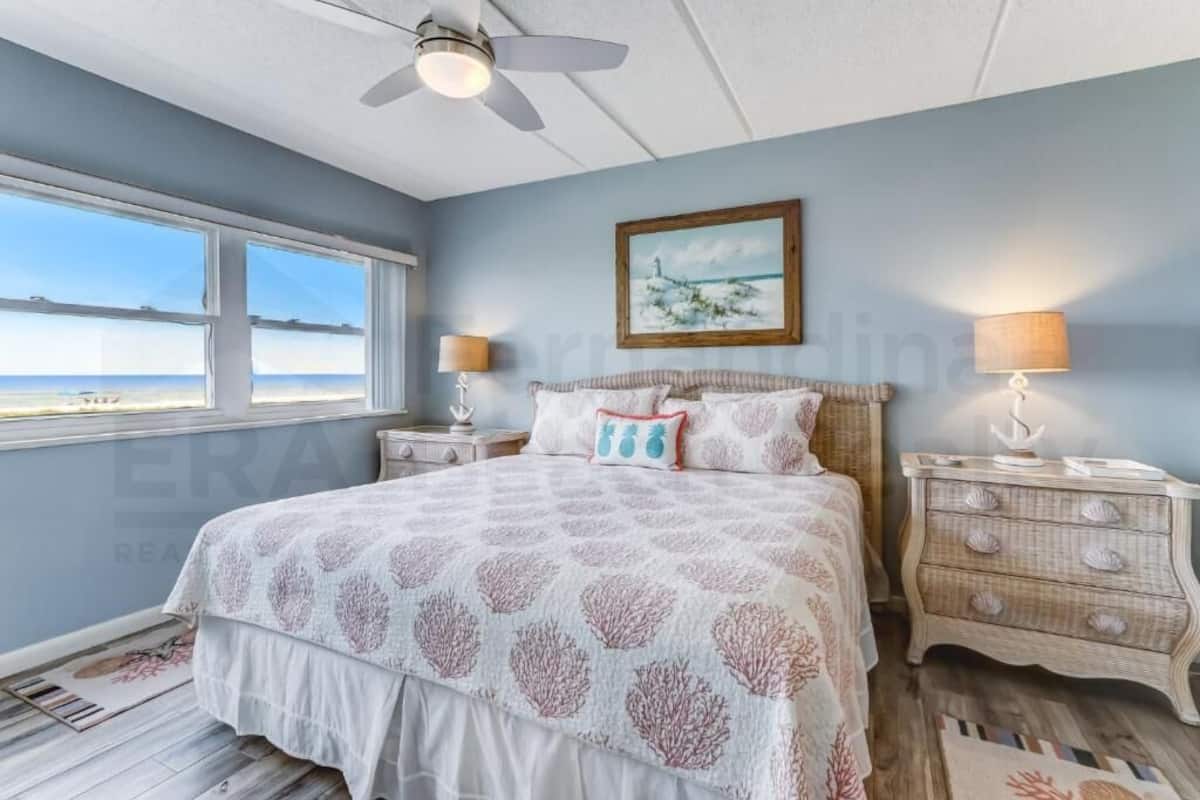 A bedroom with a bed, two nightstands, a ceiling fan, a window with a view, and a painting on the wall.