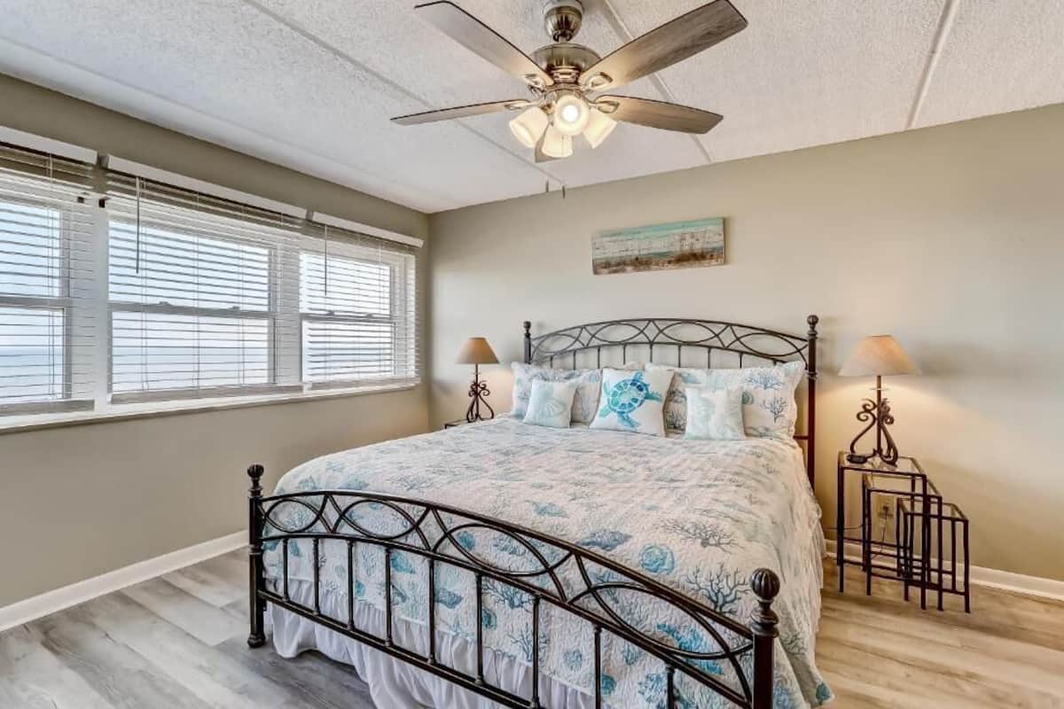 A bedroom with a bed, ceiling fan, window blinds, and a painting on the wall.