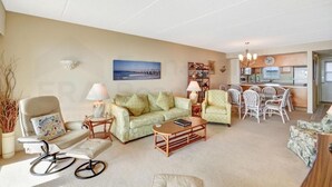 Un salon avec un canapé, un fauteuil et une table basse, un coin repas avec des chaises, et une cuisine à l’arrière-plan.