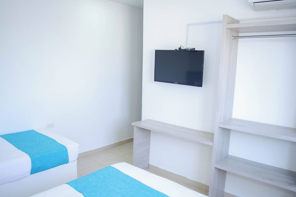A hotel room with two beds, a mounted TV, and a wooden shelf unit.