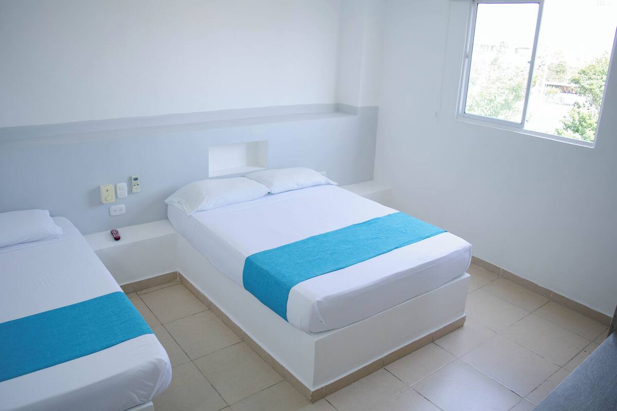 A hotel room with two beds, each with white bedding and a blue accent. There is a window providing natural light, and the room has a simple, clean design with a tiled floor.