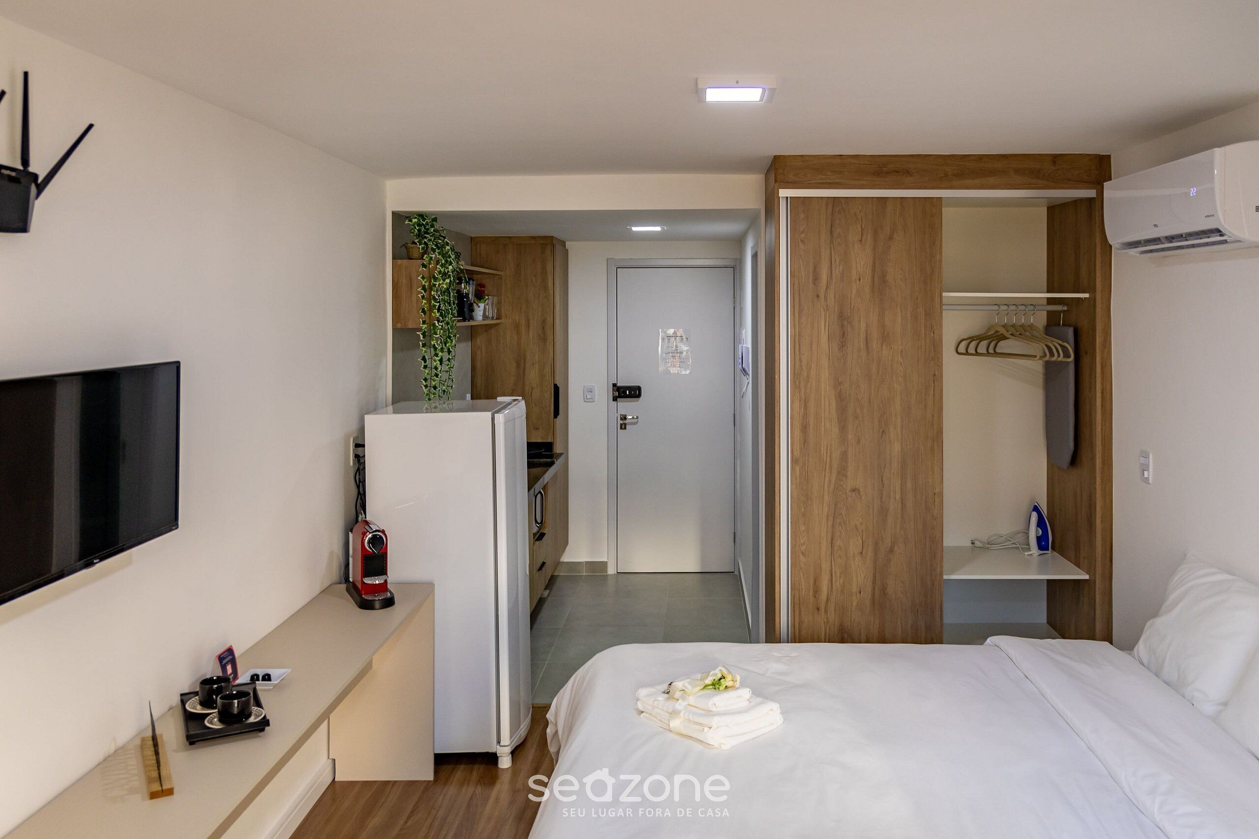 A hotel room with a bed, a TV, a desk with a coffee maker, a refrigerator, and a wardrobe.